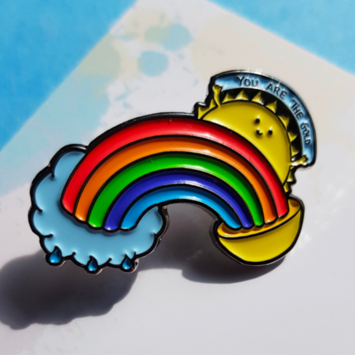 "A colorful soft enamel pin featuring a rainbow, sun, and pot of gold with a banner reading ‘You Are the Gold.’ with vibrant details, displayed on a backing card."