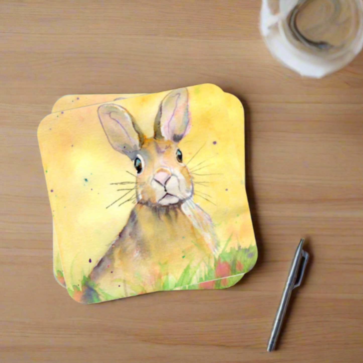 a coaster on a desk next to a pen and a cup of coffee, featuring a print of an original watercolour painting by artist Jagos Art of a startled yet cheeky looking hare sitting in the grass with a vibrant yellow background