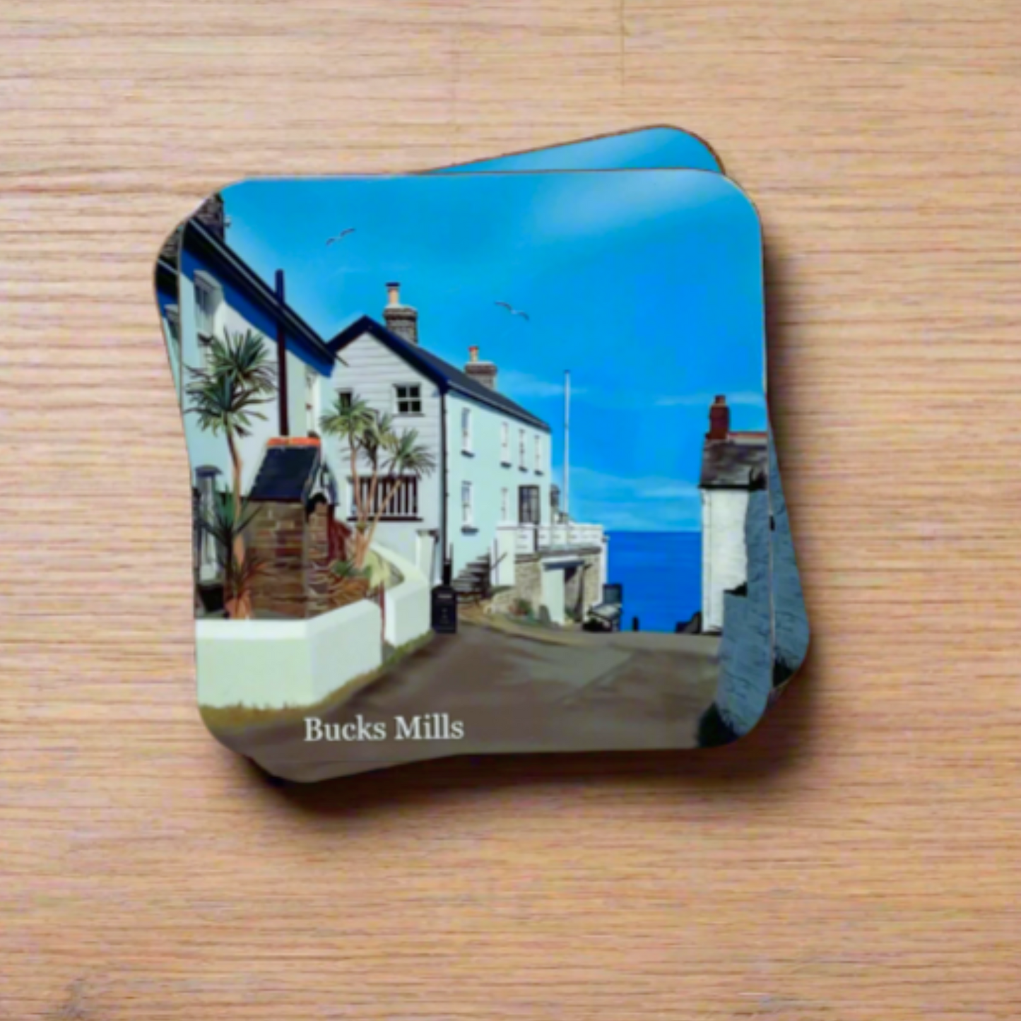 "A square glossy coaster on a desk, featuring a digital illustration of Bucks Mills by artist Jagos Art. The artwork showcases historic buildings, and the start of the journey down to the beach. Printed using the sublimation process."