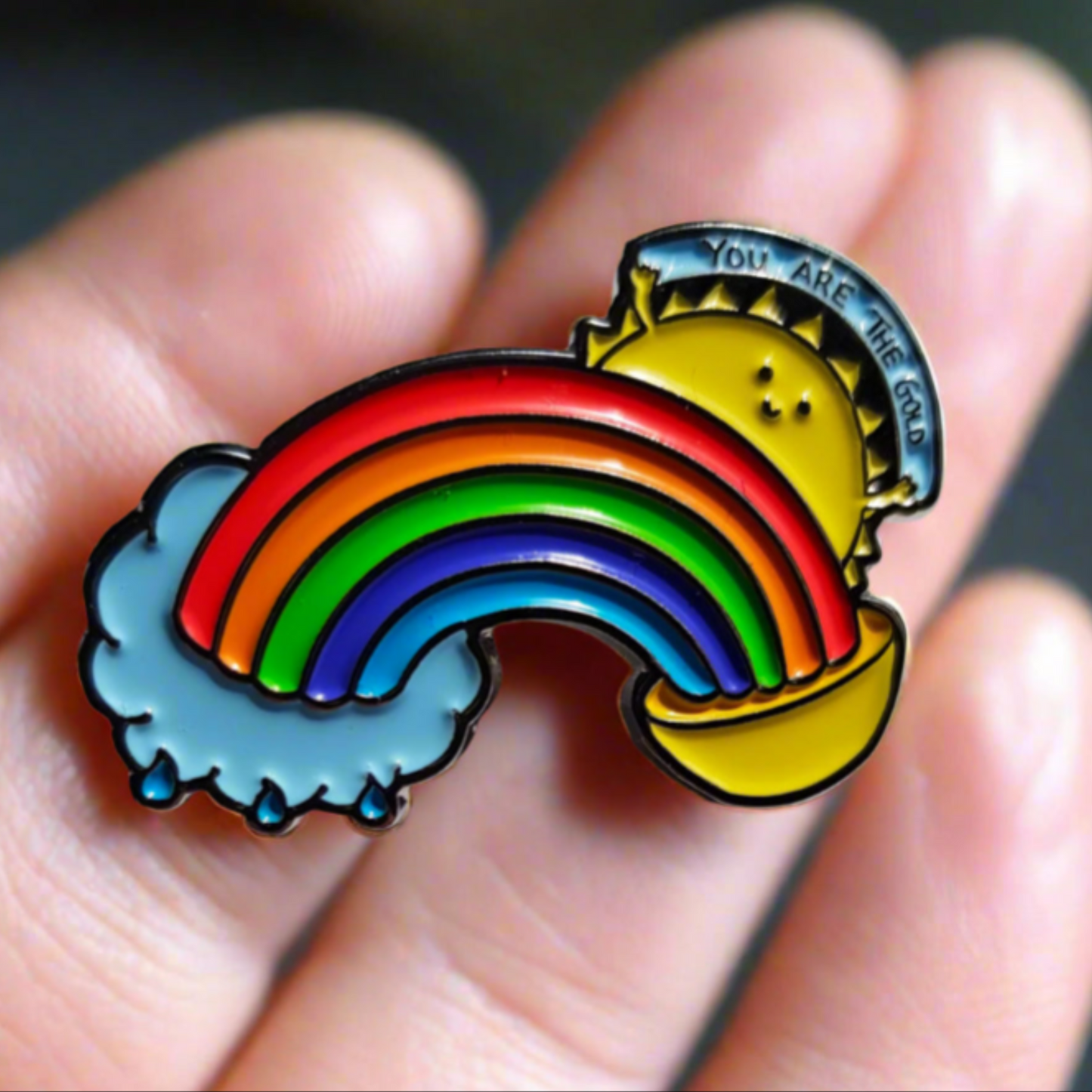 "A close-up of a ‘You Are the Gold’ enamel pin, held delicately on some fingertips. The rainbow and sun design symbolizes self-worth, positivity, and encouragement."