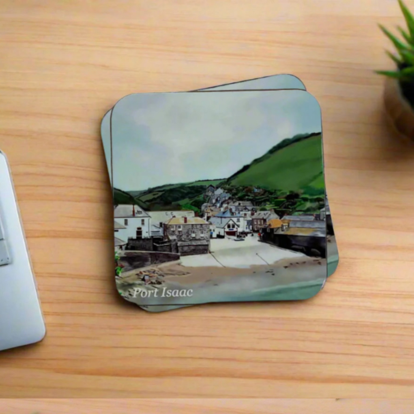 "A square glossy coaster on a desk, featuring a digital illustration of Port Isaac by artist Jagos Art. The artwork showcases harbour shops, the beach slipway, historic buildings, and scenic fields in the background. Printed using the sublimation process."