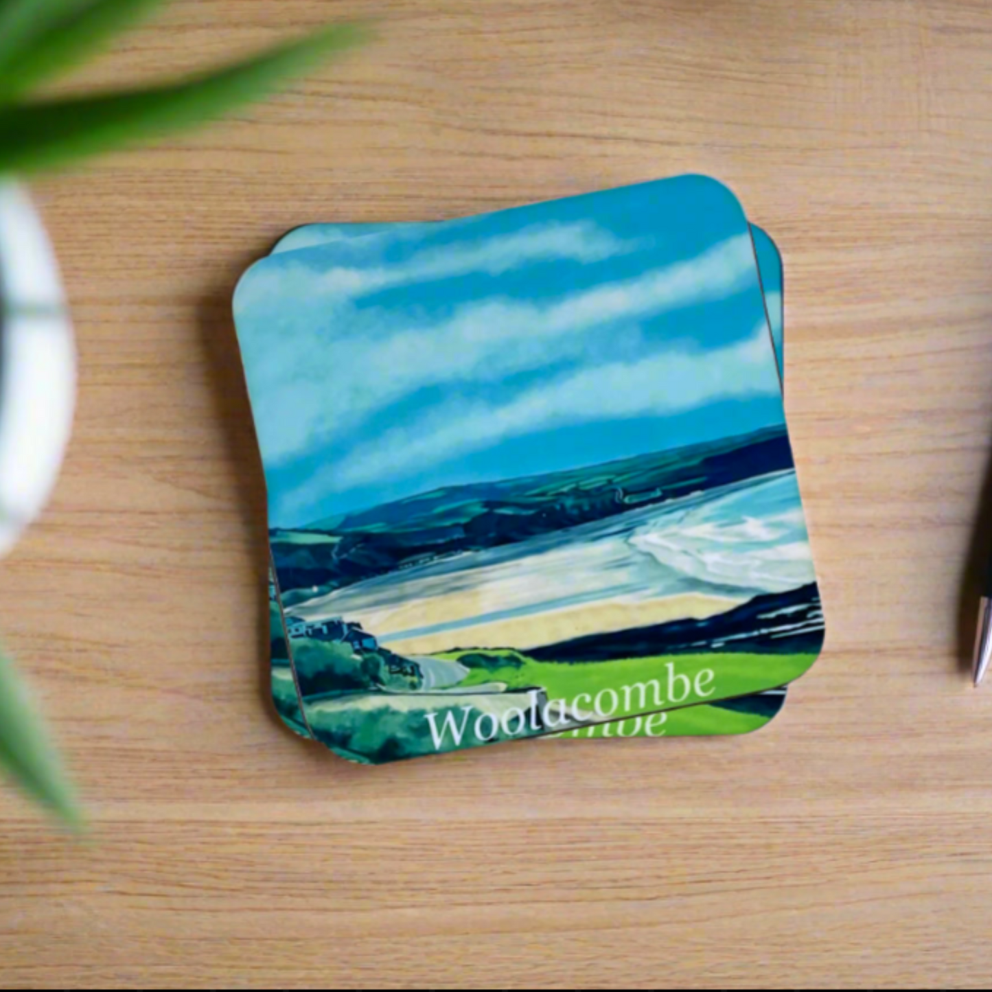 "A square glossy coaster on a desk, featuring a digital illustration of Woolacombe by artist Jagos Art. The artwork showcases the beach and the field landscape behind. Printed using the sublimation process