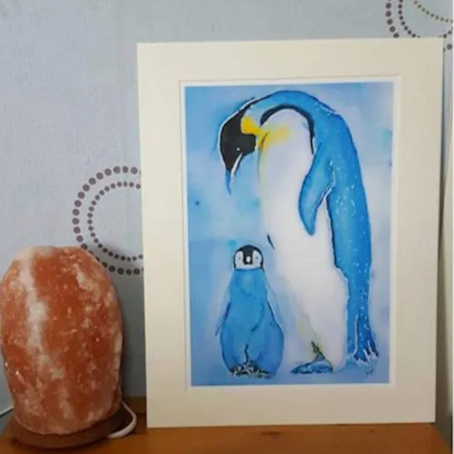An A4 art print featuring an original watercolour painting by artist Jagos Art. The artwork showcases a tender moment between a mother and baby penguin, with the baby standing at the mother's feet while she looks down lovingly. Painted in blue-grey, white, and soft yellow tones. Printed on archival paper for lasting vibrancy."