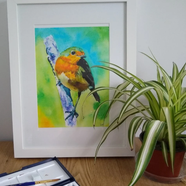 "An framed A4 robin art print on a table, surrounded by a plant and brush on a watercolour palette. The detailed brushstrokes and natural colours highlight the beauty of this handmade wildlife portrait print.