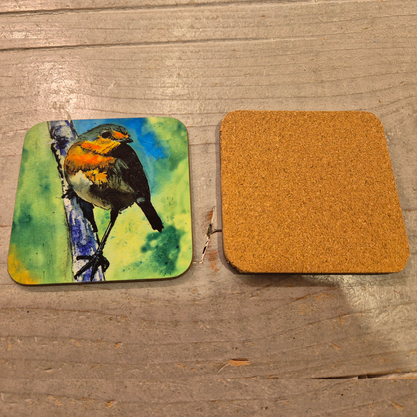 "A pair of square coasters on a wooden surface, one facing upwards featuring a print of an original watercolour painting by artist Jagos Art, the second facing downwards showing the cork backing. The artwork depicts a vibrant robin perched on a branch against a lush green background."