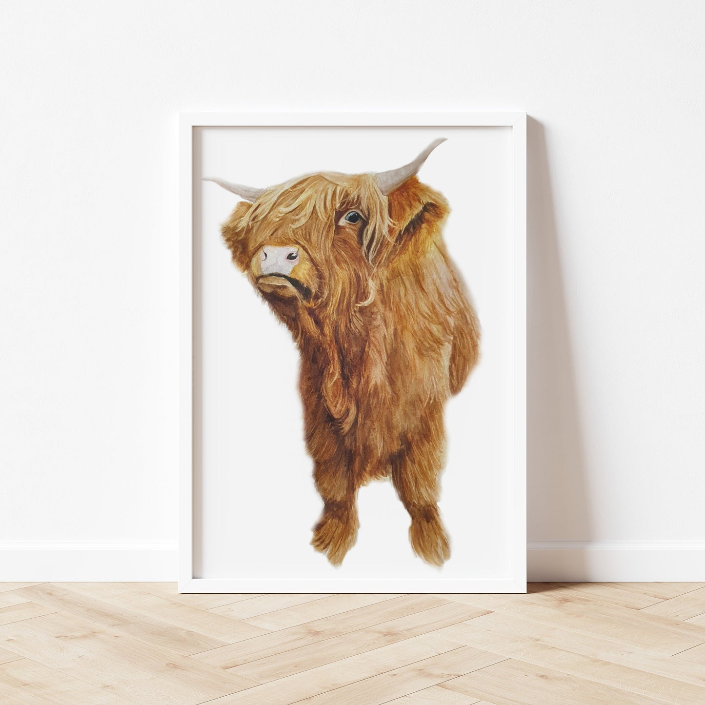 "An A4 art print, in a white frame stood against a wall, featuring an original watercolour painting by artist Jagos Art. The artwork showcases a young Highland cow standing forward-facing, its shaggy auburn forelock covering most of its face except for one expressive eye. Printed on archival paper with a crisp white background."