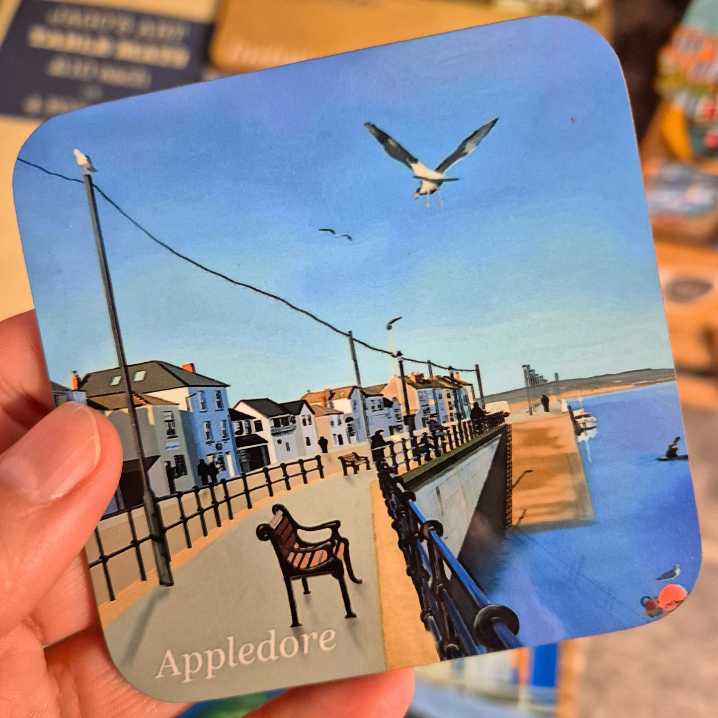 Appledore coaster by Jagos Art, with blue sky, blue water, seagulls, shops and benches, held in hand