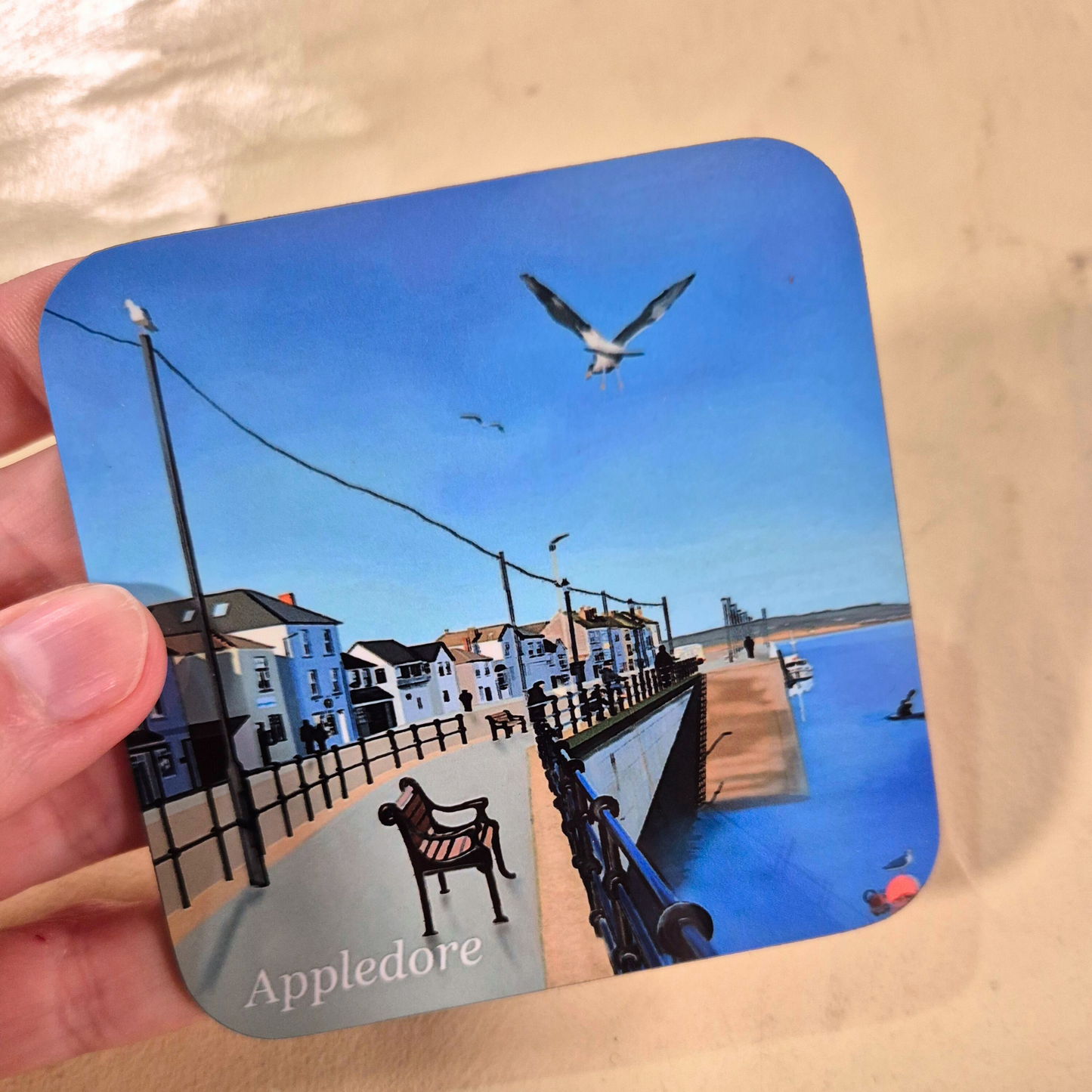 Appledore coaster by Jagos Art blue sky, blue water, seagull, bench, shops held in hand
