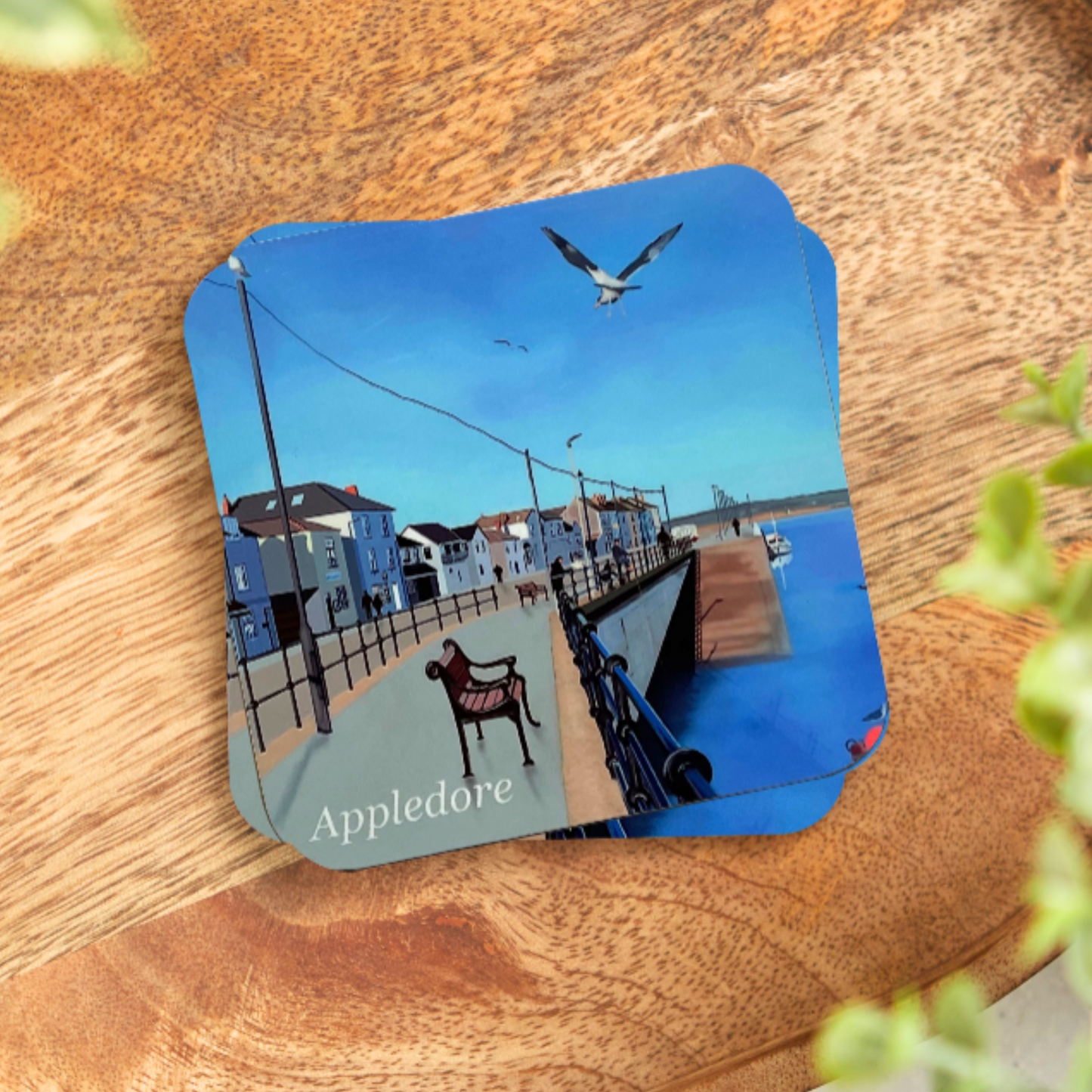 Appledore coaster by Jagos Art blue sky, blue water, bench, seagull and shops, placed on a desk