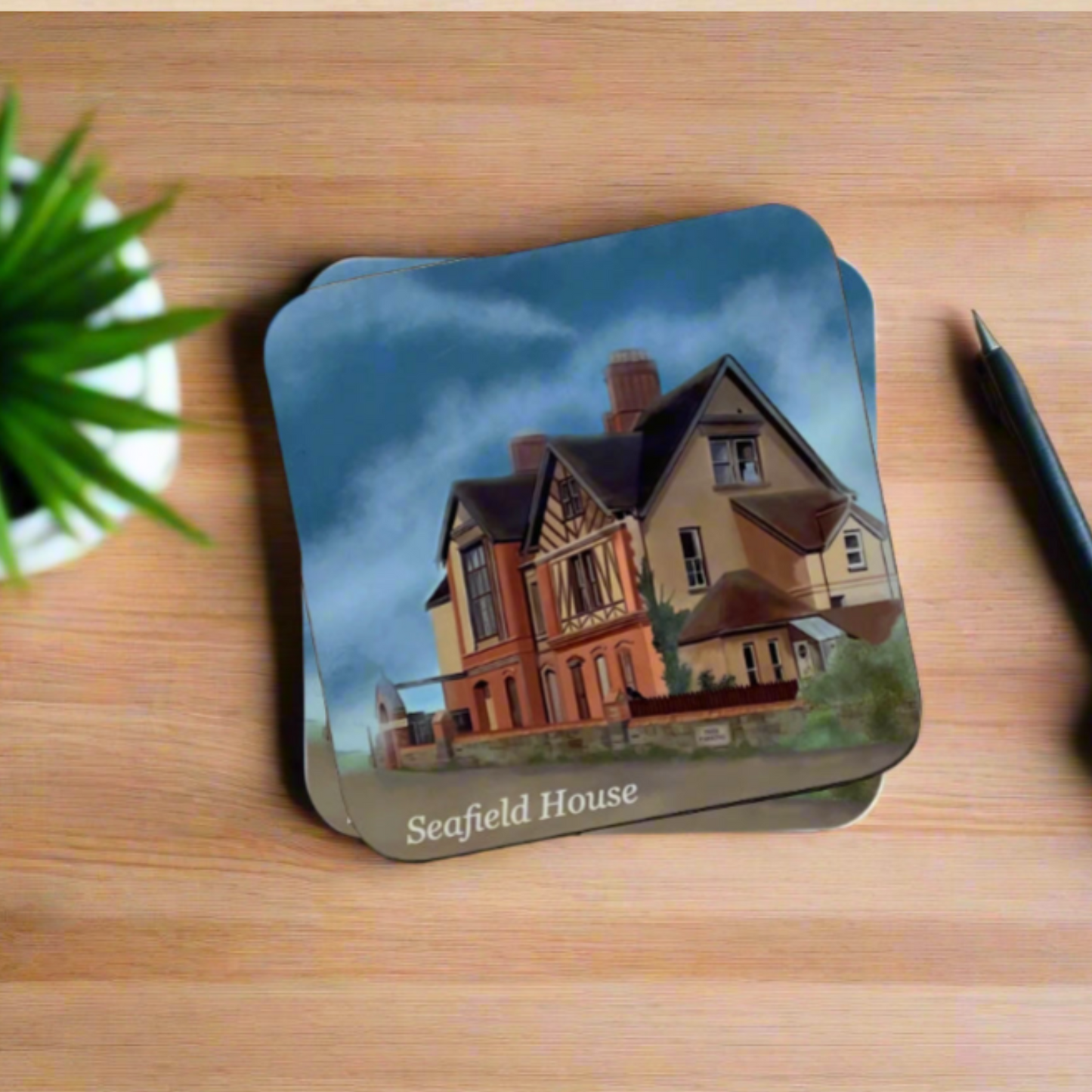 Seafield House and cloudy skies coaster by Jagos Art on a desk with pen and plant