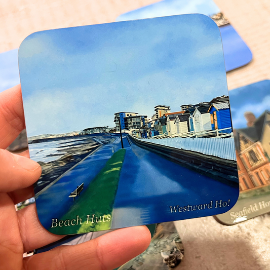 "Hand holding a square coaster featuring a hand-drawn digital painting of the beach huts at Westward Ho!, North Devon. The artwork captures the colorful huts, the scenic promenade, and the coastal walkway under a bright blue sky. The coaster has a glossy finish and is handmade using the sublimation process."