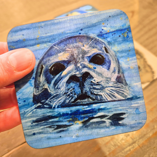 Hand holding a coaster featuring a print of an original watercolour painting by the artist Jagos Art of a seal curiously poking his head out of the water
