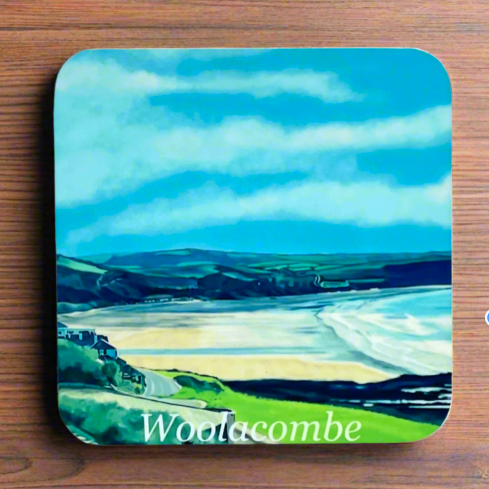 "A square glossy coaster on a desk, featuring a digital illustration of Woolacombe by artist Jagos Art. The artwork showcases the beach and the field landscape behind. Printed using the sublimation process