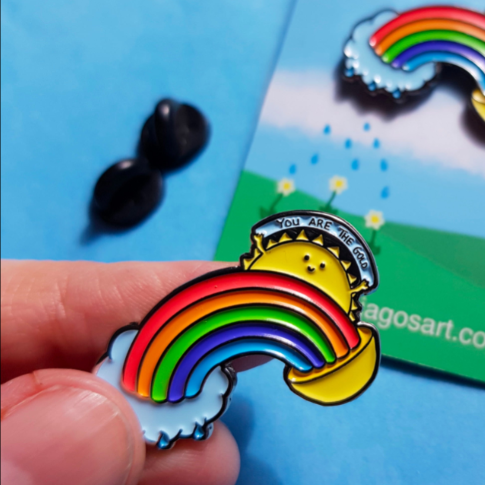 "A close-up of a ‘You Are the Gold’ enamel pin, held delicately between fingertips. The rainbow and sun design symbolizes self-worth, positivity, and encouragement." In the background is the backing card featuring the pin and on the card are raindrops shown falling from the little cloud, watering some flowers