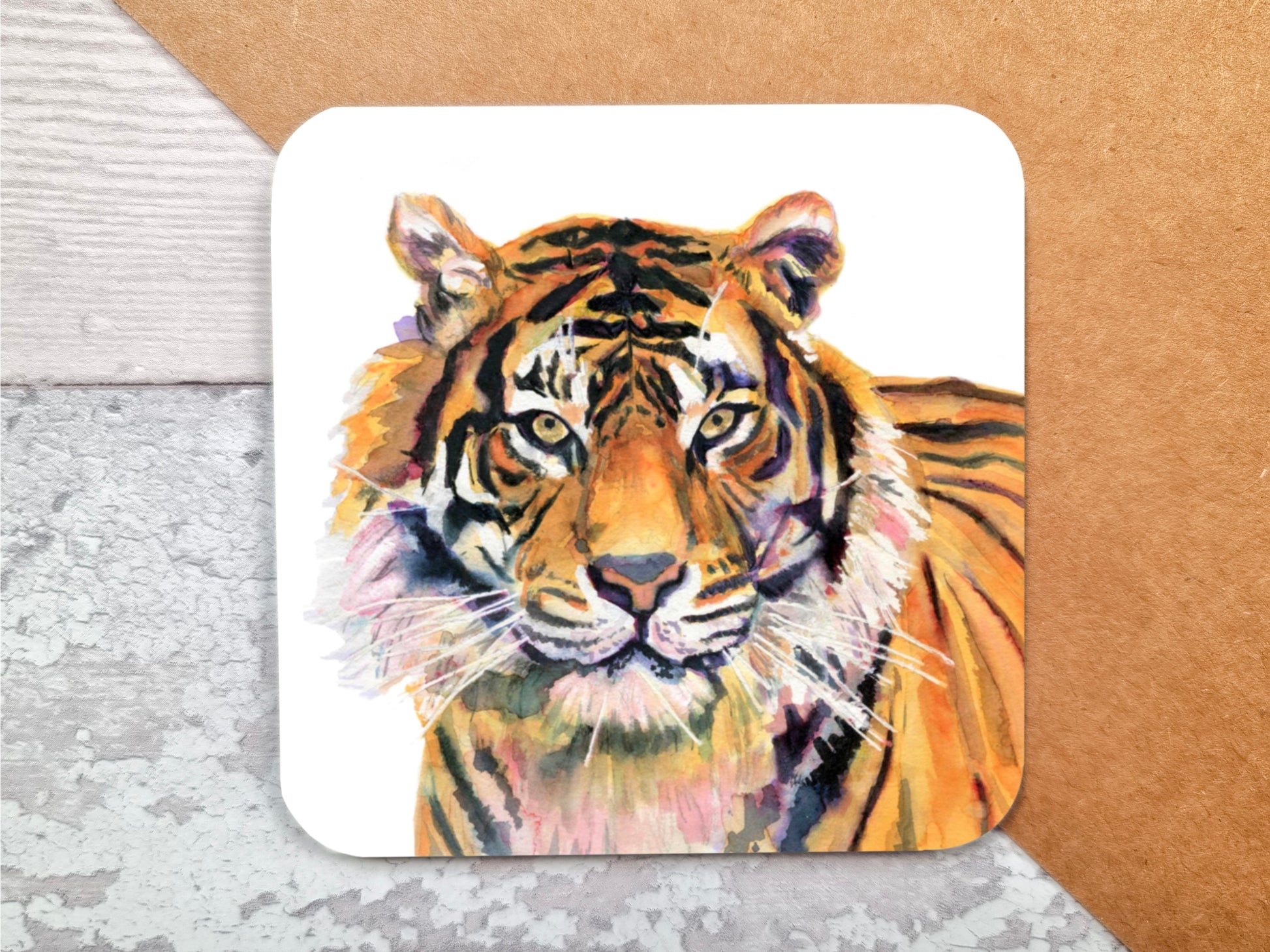 A coaster featuring a print of an art print of an original watercolour painting of a stunning Tiger looking right at the viewer with the primary colours being orange with purple tones