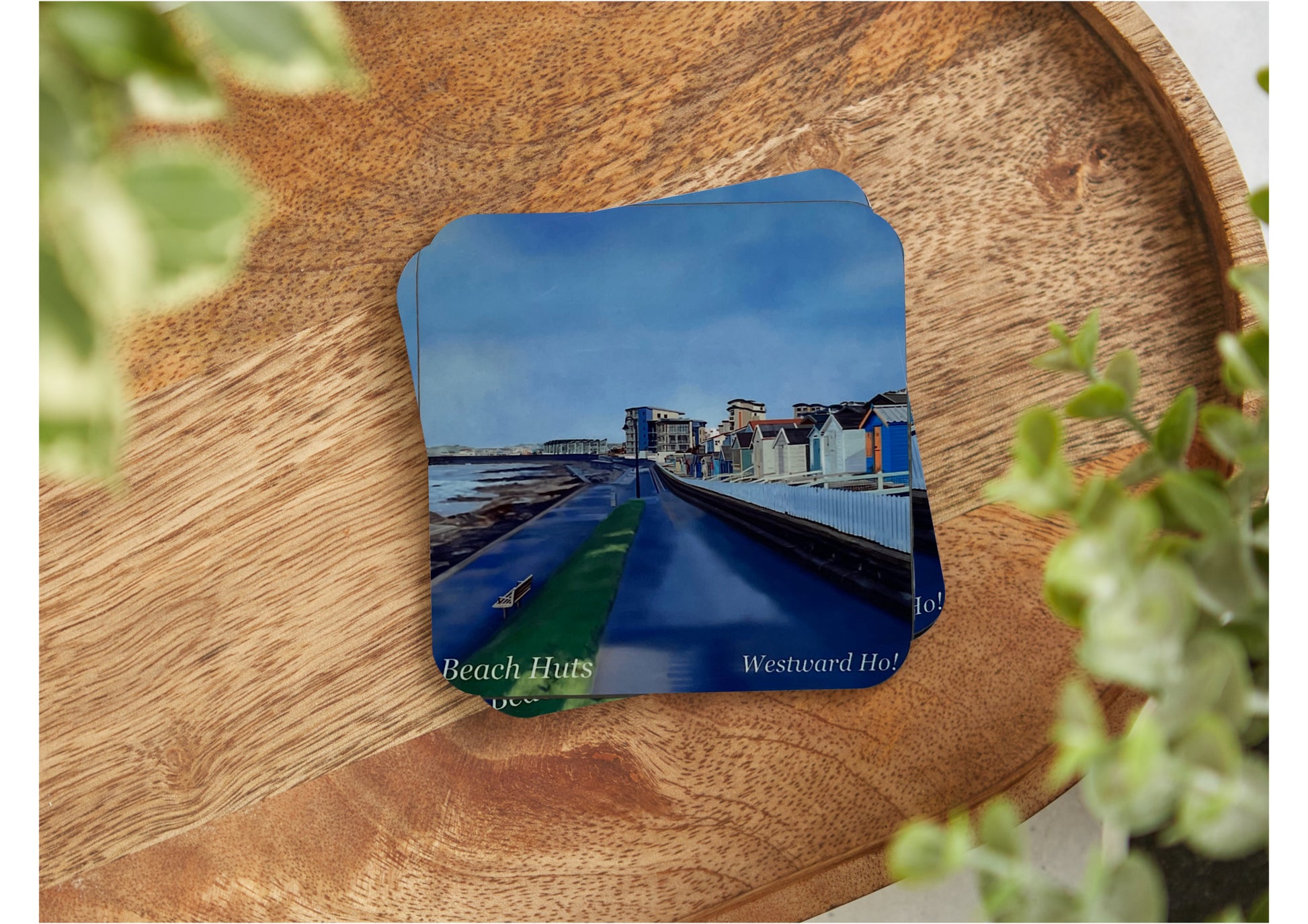 A table coaster featuring a print of Westward Ho in North Devon showing the walkway beside the sea, the row of beach huts and some of the village in the distance