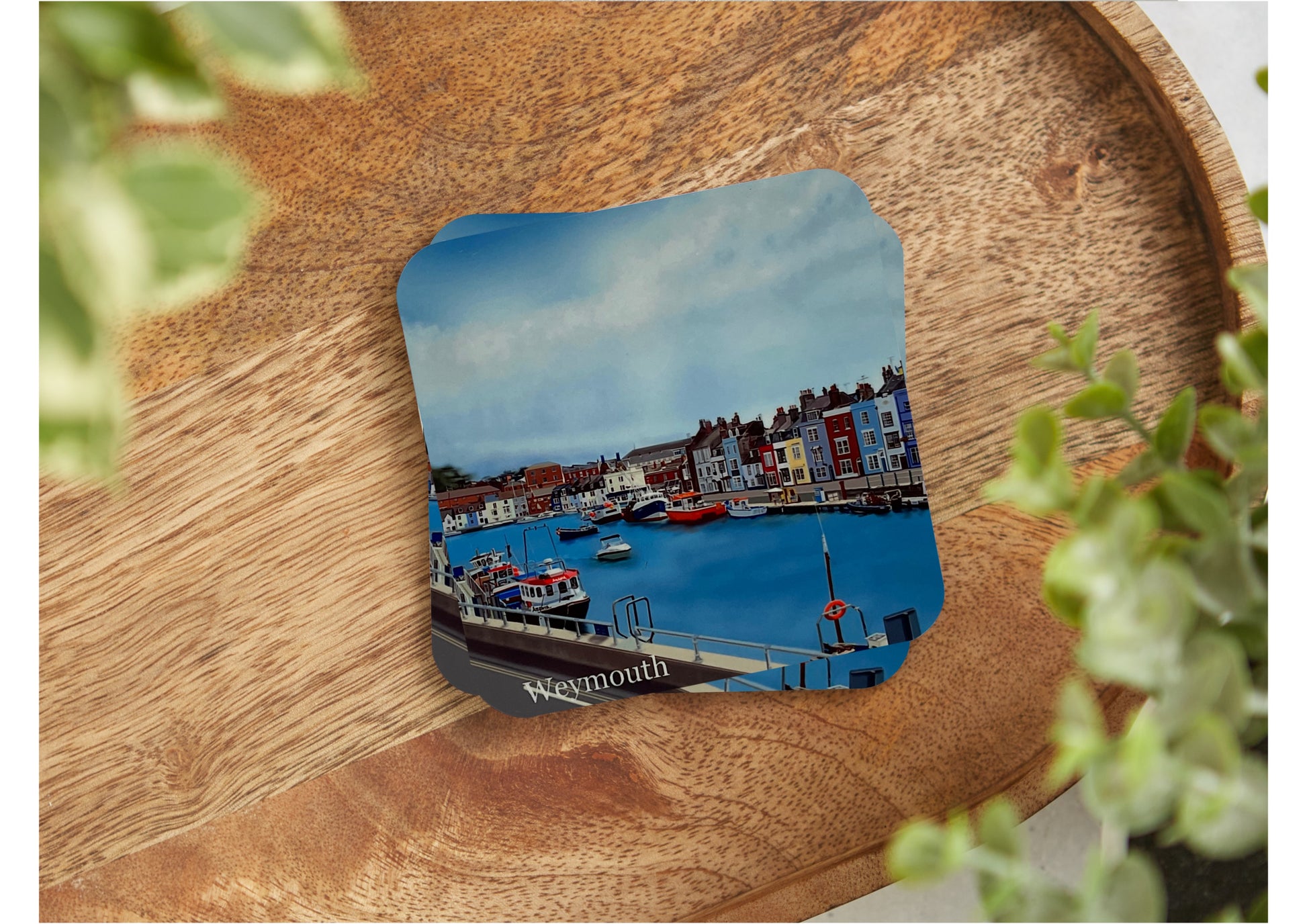 A table coaster featuring a print of Weymouth harbour showing the river with several boats moored up and the colourful buildings