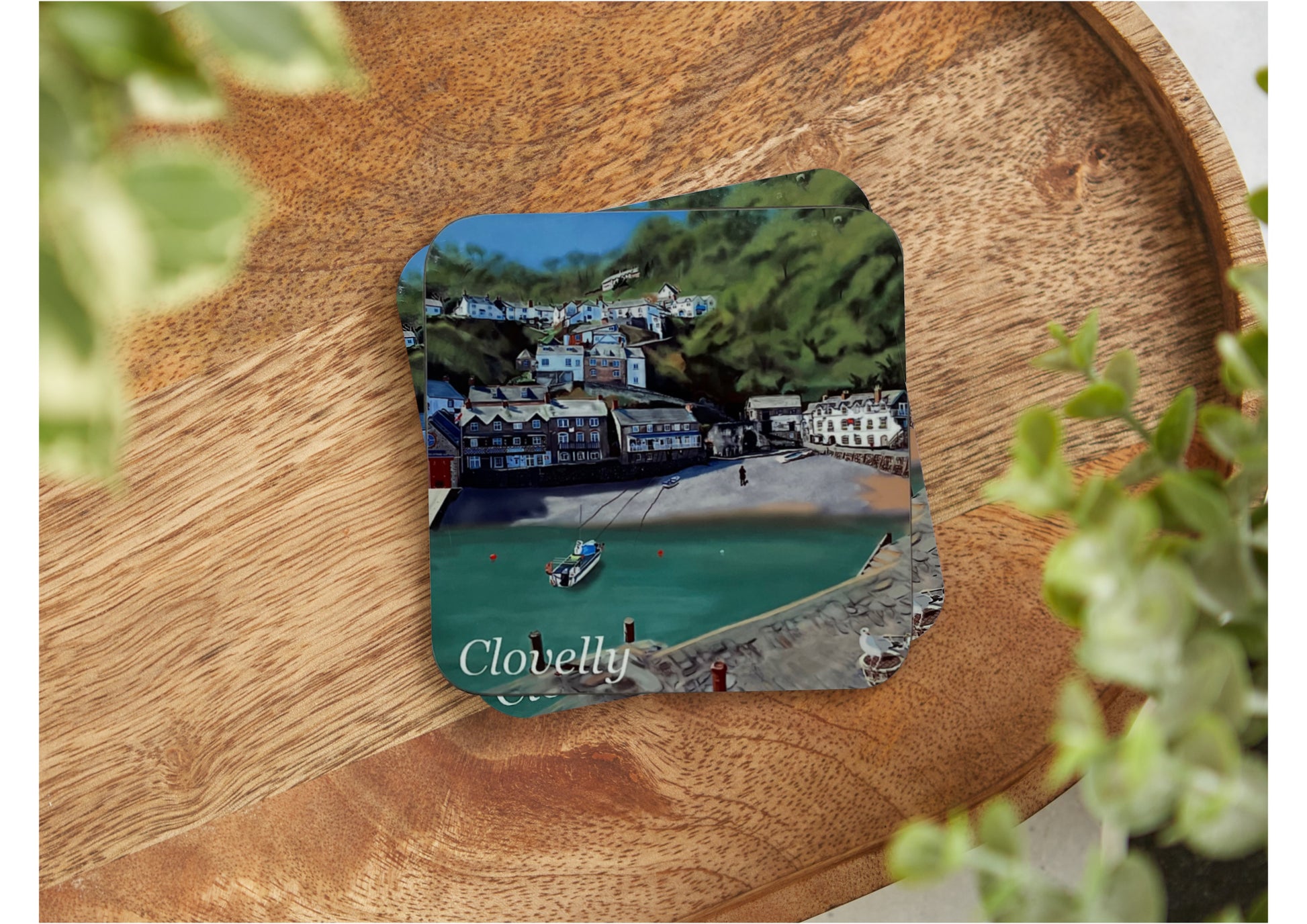 A table coaster handmade by Jagos Art featuring a print by Jagos Art of Clovelly Harbour in North Devon showing the buildings from top to bottom, the hotel and the small beach with green being the primary colour