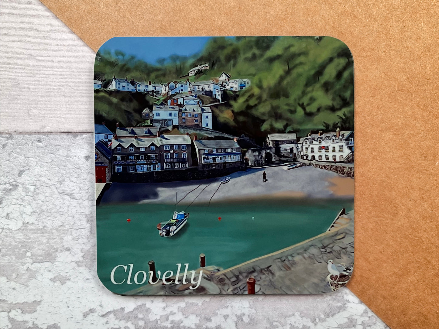 A table coaster handmade by Jagos Art featuring a print by Jagos Art of Clovelly Harbour in North Devon showing the village, the small beach, a small fishing boat and the hotel 