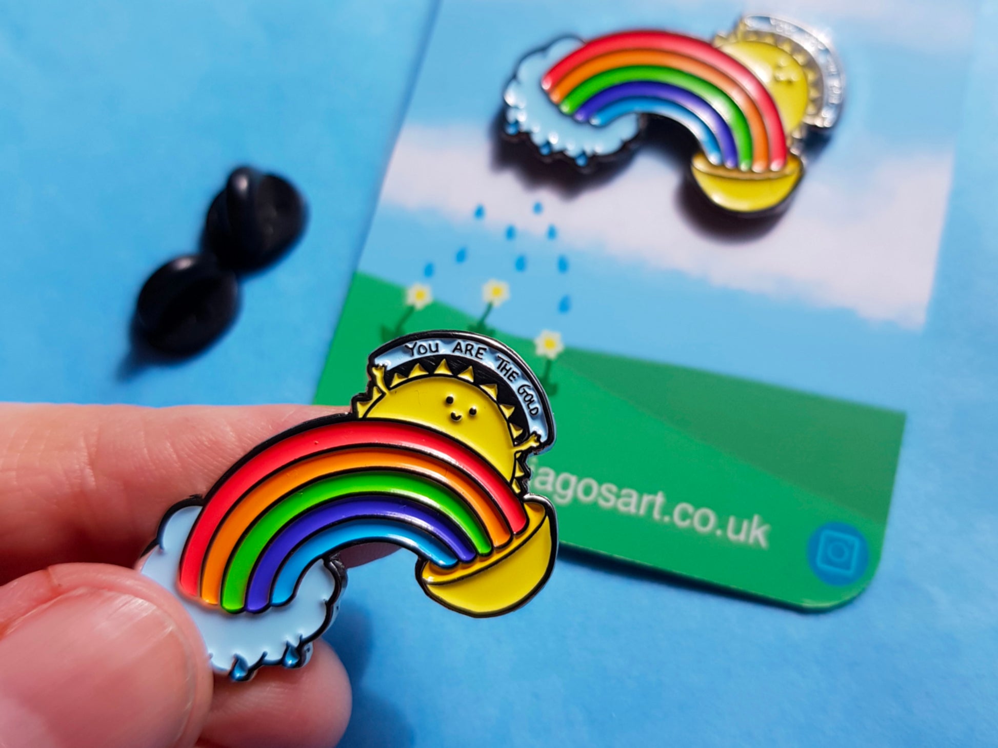 A photo of an enamel pin of a rainbow coming our of a small rain cloud at one end and a pot of gold at the other and from above the rainbow a sun is shining and the sun has a little smiling face and two small hands holding up a sign saying you are the gold