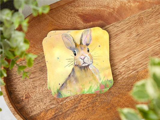 A handmade coaster featuring an original watercolour painting by Jagos Art of a Hare Bunny sitting in the grass and the flowers with an inquistive look on its face looking right at the viewer with a yellow background