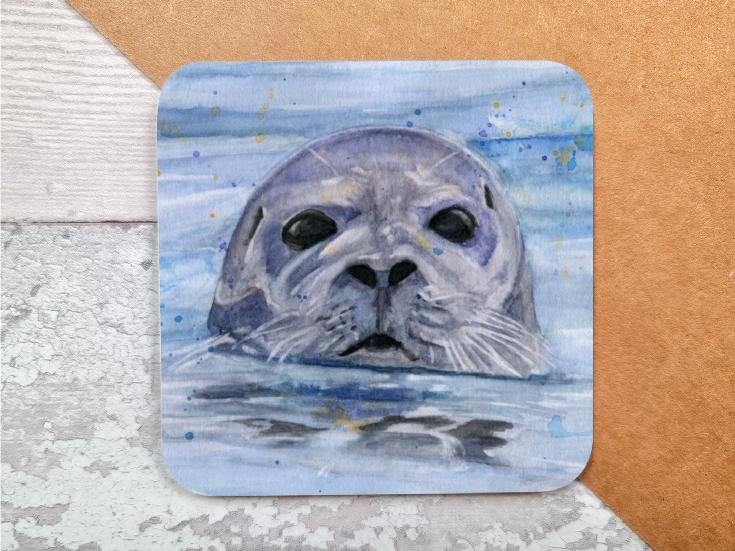 A coaster featuring a print of an original watercolour painting of a Seal popping his head up out of the water with the primary colours being blue and grey