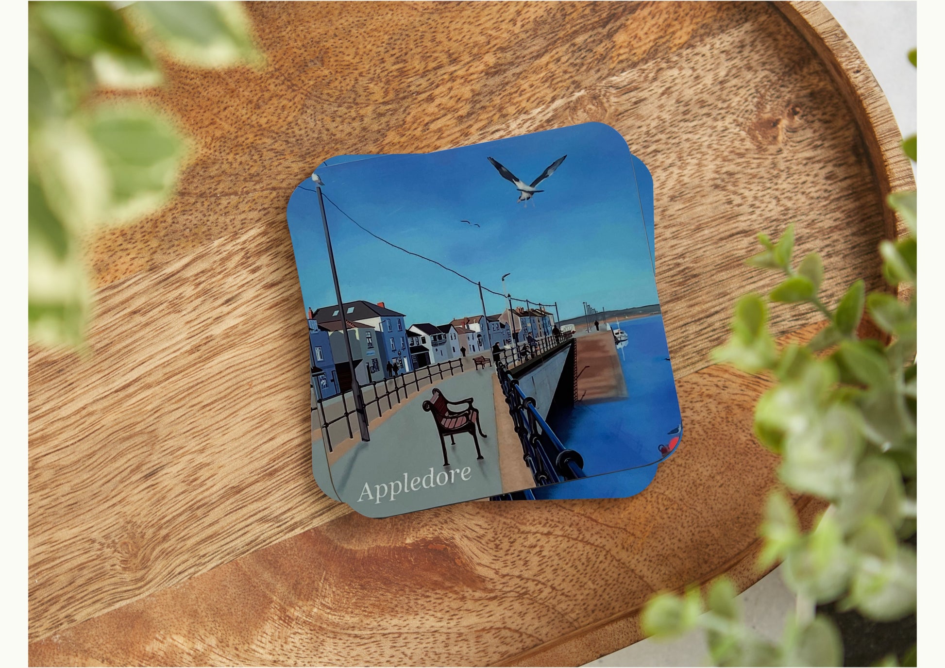 A table coaster featuring a print of Appledore Quay in North Devon by Jagos Art with blue skies, a blue river, buildings and seagulls