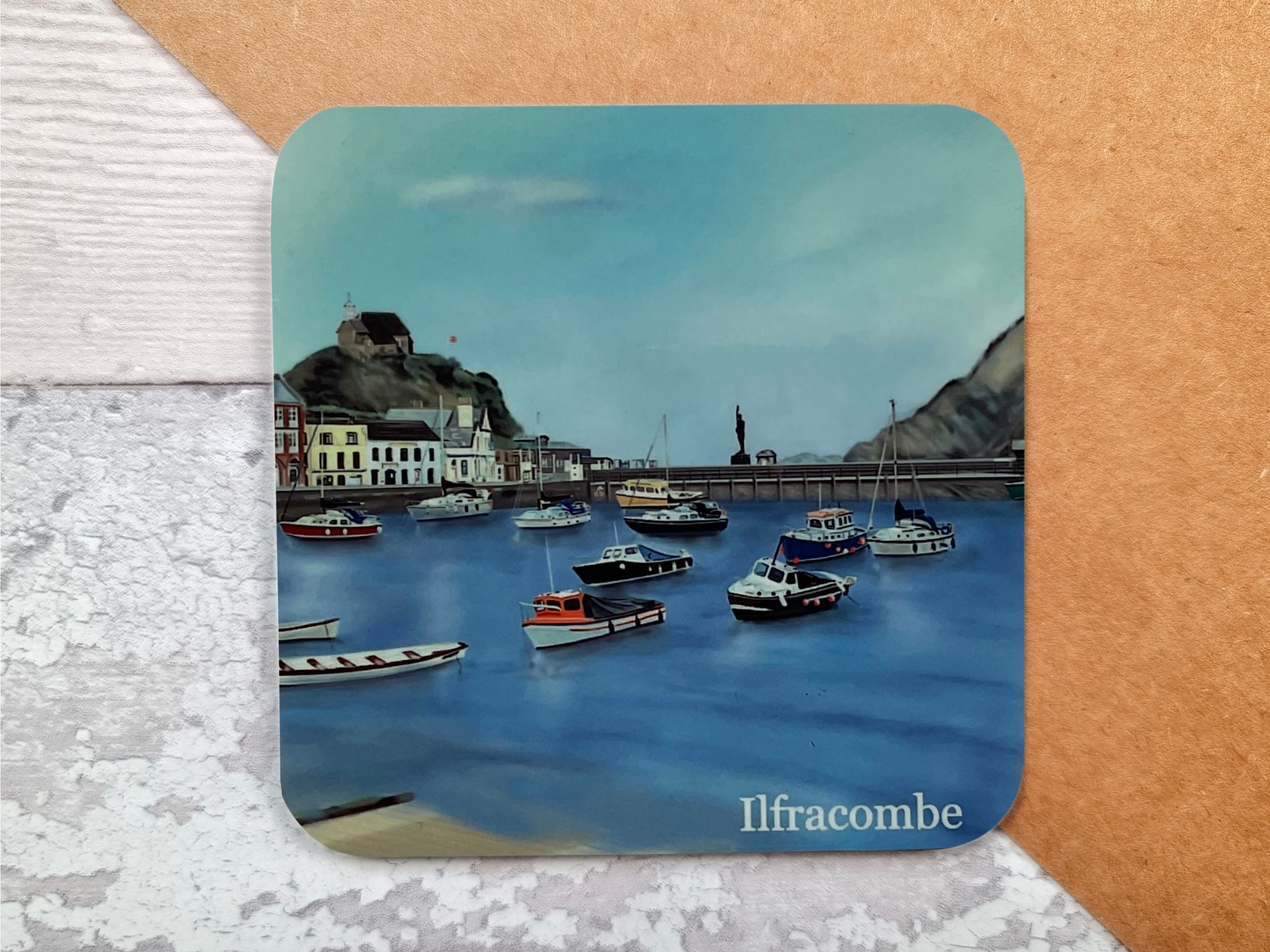 A table coaster featuring a print of Illfracombe Harbour showing some of the buildings, several fishing boats moored up, the verity statue in the distance and the church on the hill