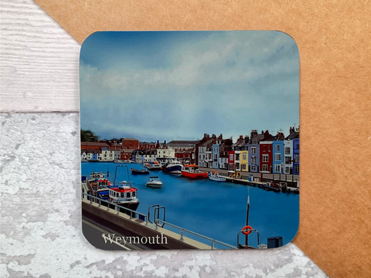 A table coaster featuring a print of Weymouth Harbour showing the river, several boats moored up and the colourful buildings