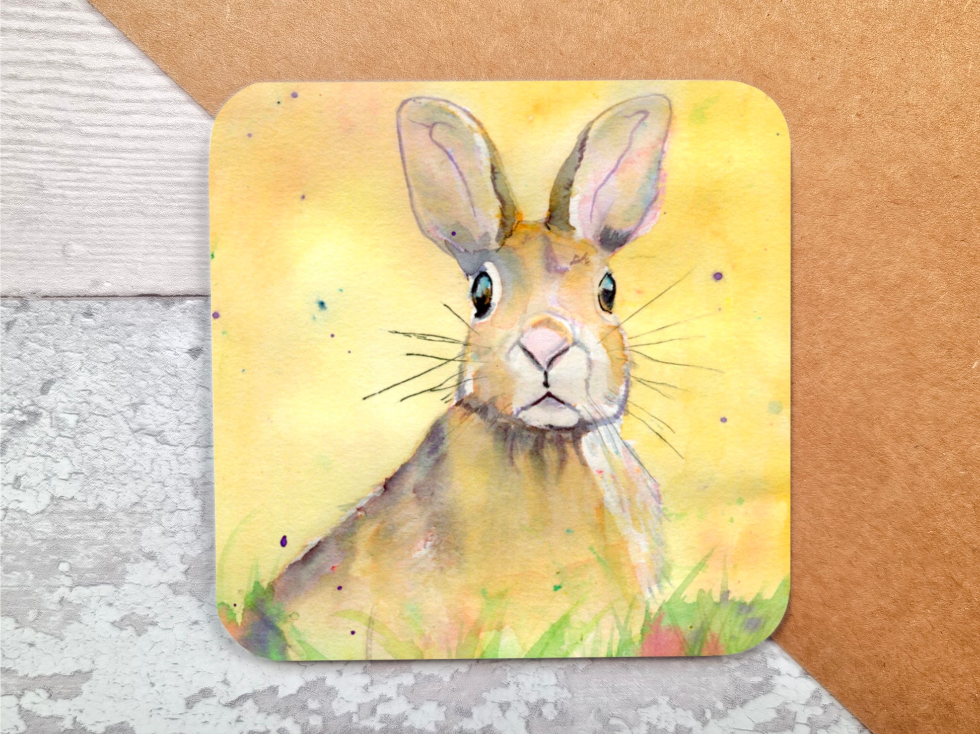 A handmade coaster featuring an original watercolour painting by Jagos Art of a Hare Bunny sitting in the grass and the flowers with an inquistive look on its face looking right at the viewer with a yellow background