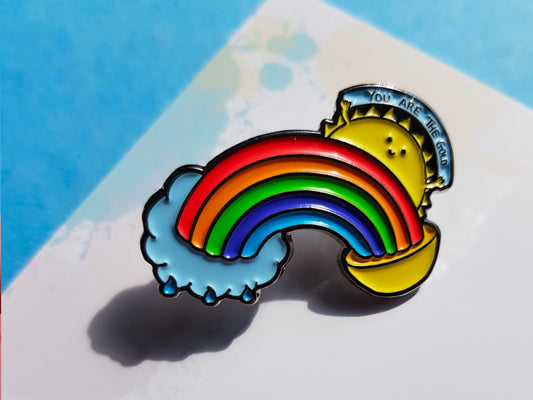 A photo of an enamel pin of a rainbow coming our of a small rain cloud at one end and a pot of gold at the other and from above the rainbow a sun is shining and the sun has a little smiling face and two small hands holding up a sign saying you are the gold