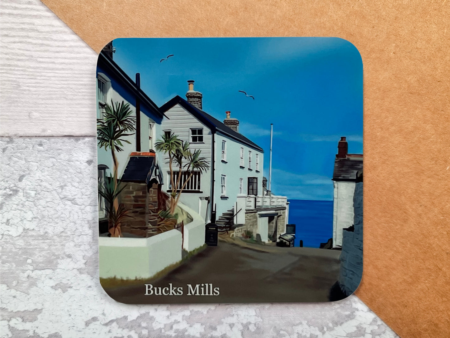 A table coaster handmade by Jagos Art featuring a print of Bucks Mills in North Devon showing the buildings at the top of the path to the beach and the sea in the background with a couple of seagulls flying overhead